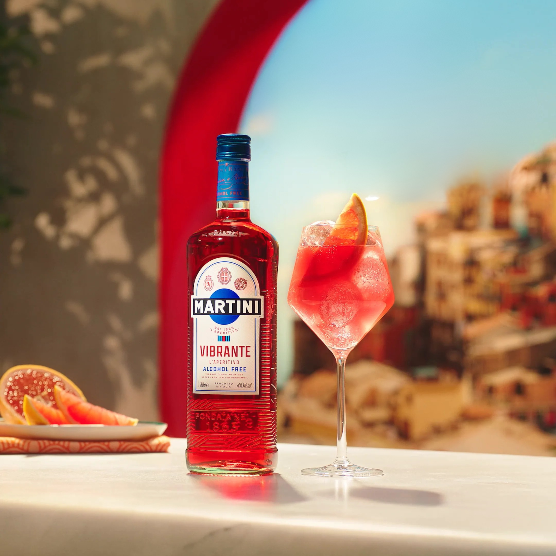 Bottle of Martini Vibrante alcohol-free liqueur with a glass of pink cocktail on a table with a scenic background.