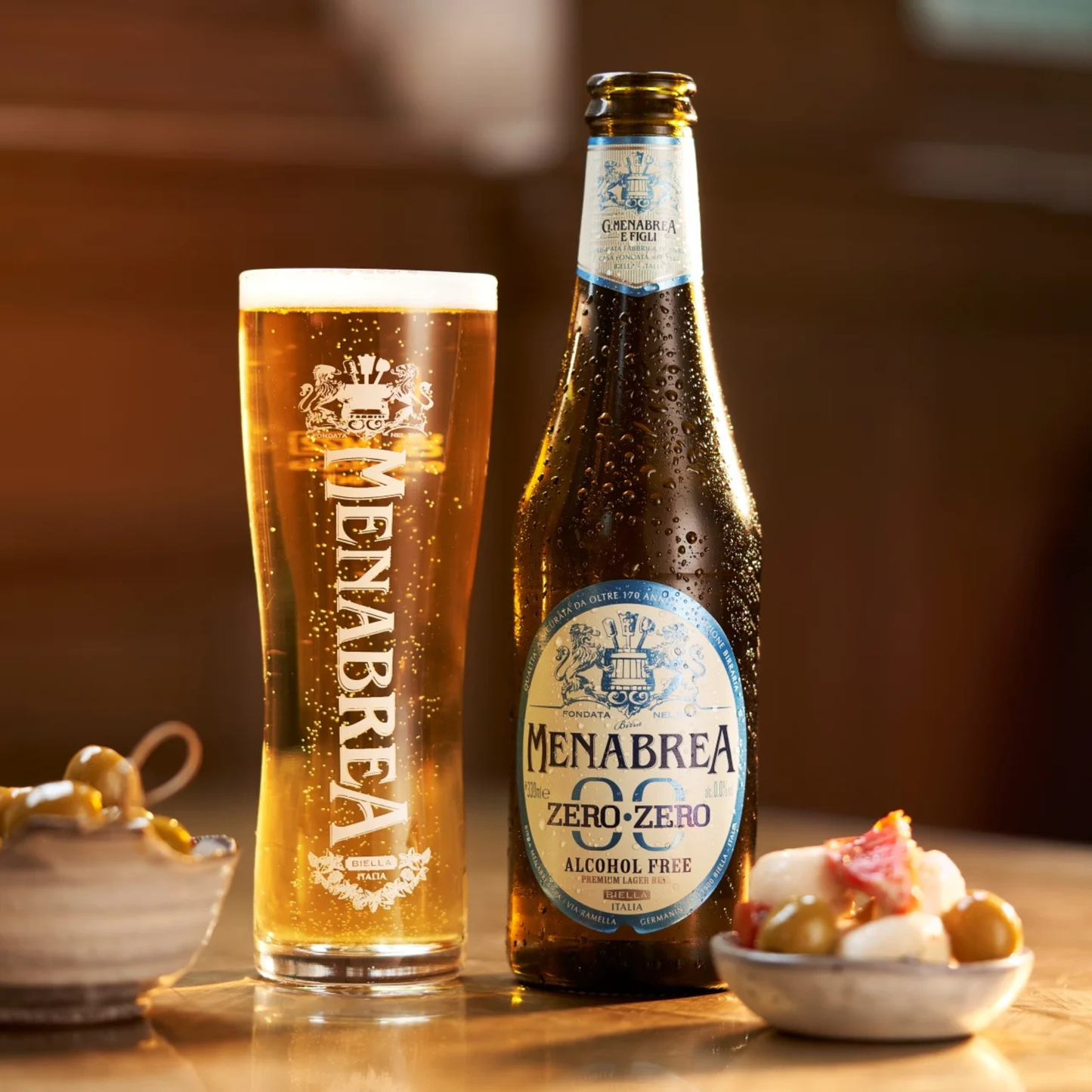 Menabrea alcohol-free beer bottle and glass on a wooden table with snacks.