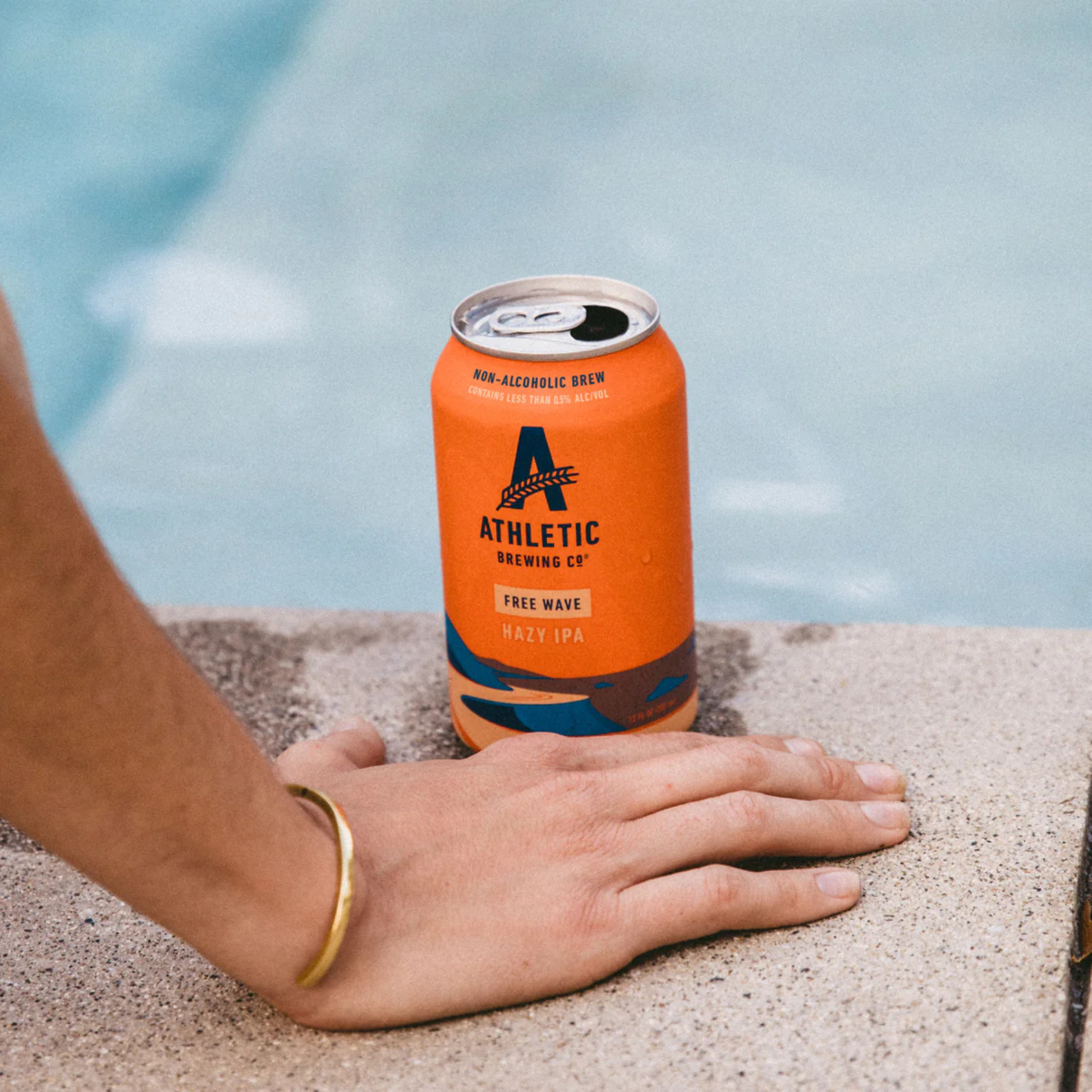 Orange can of Athletic Brewing Co. by a poolside with a hand resting on the ledge.