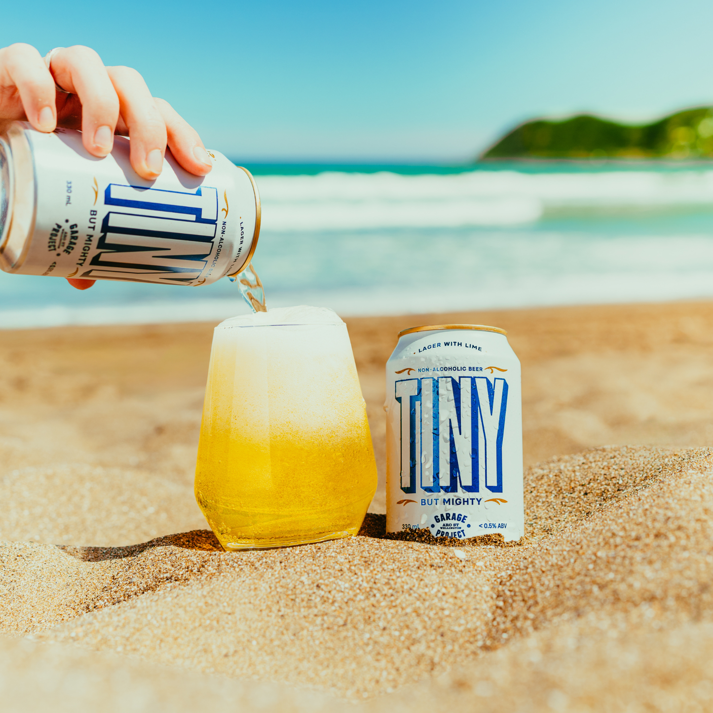 Person pouring a can of TINY beer into a glass on a sandy beach with ocean view.