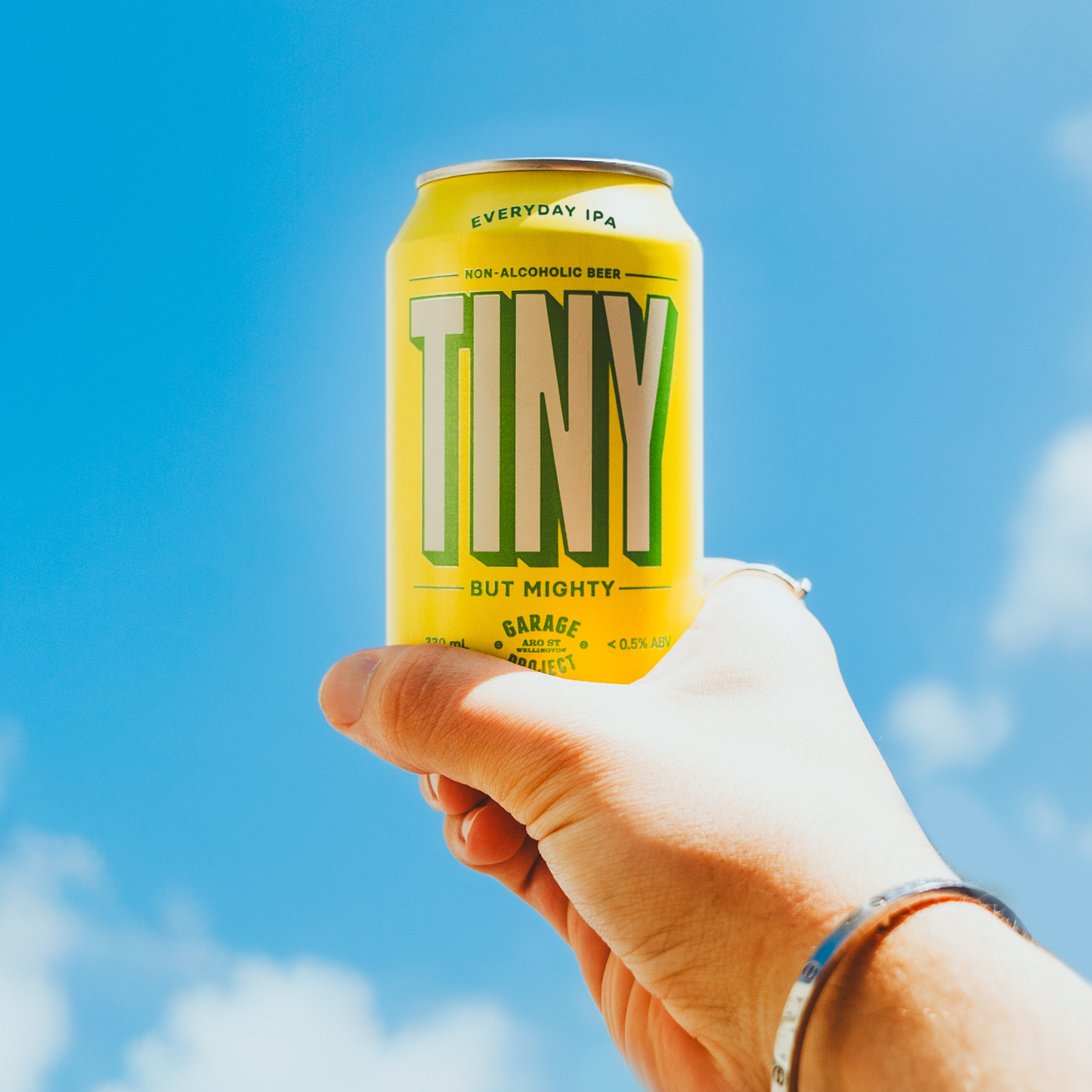 Hand holding a can of 'Tiny' non-alcoholic beer against a blue sky.