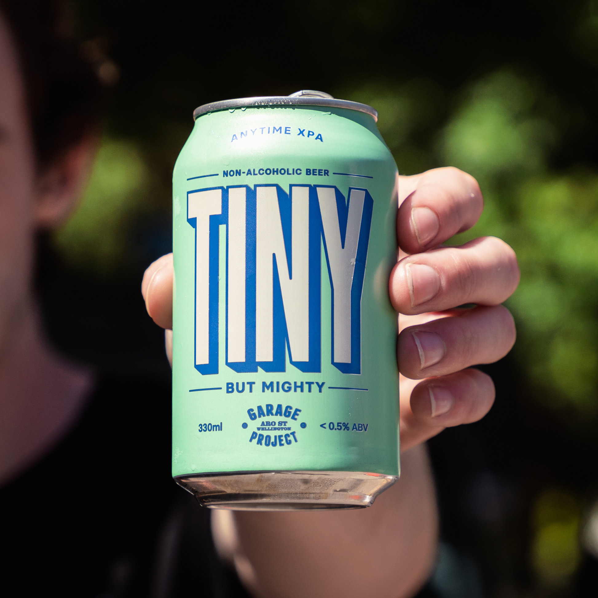 Hand holding a can of 'TINY' non-alcoholic beer with a blurred green background