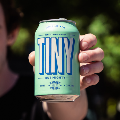 Hand holding a can of 'TINY' non-alcoholic beer with a blurred green background