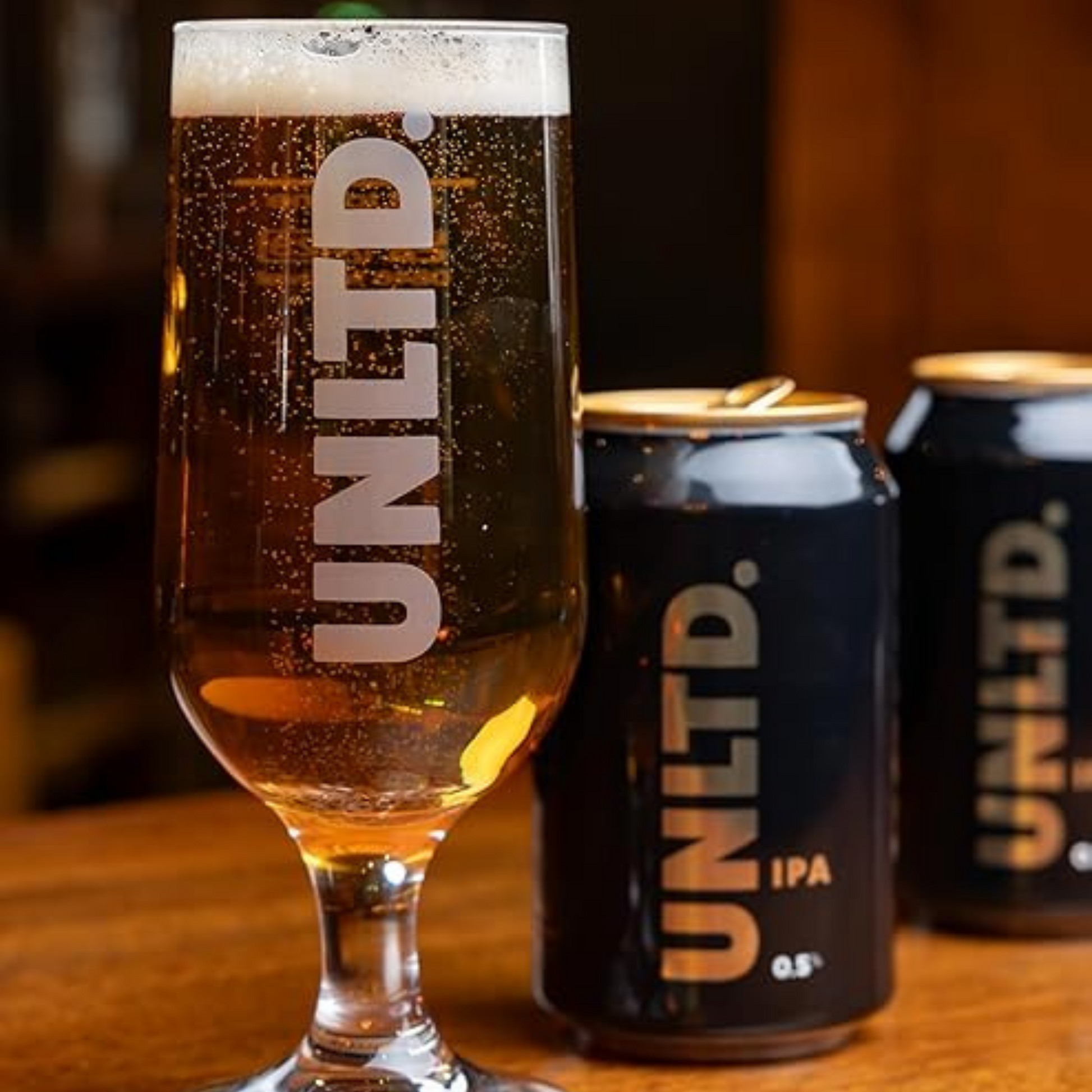 UNLTD. IPA glass and cans on a wooden surface with a blurred background